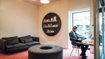 A modern room with a black sofa, a round ottoman, and a desk with a computer. There is a bookshelf with books and a large window.
