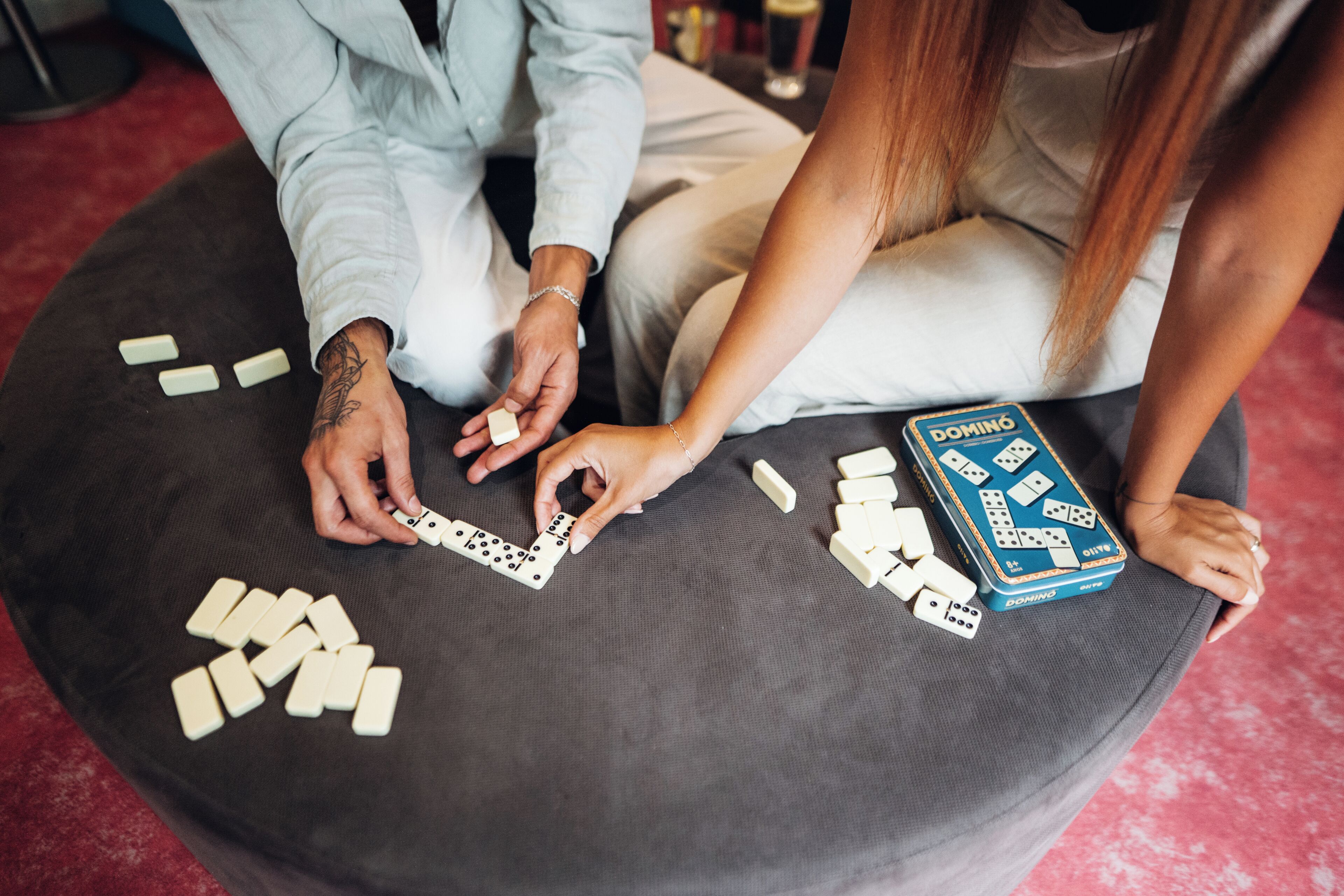 Une personne joue aux dominos sur un pouf rond, avec un jeu de dominos à portée de main.