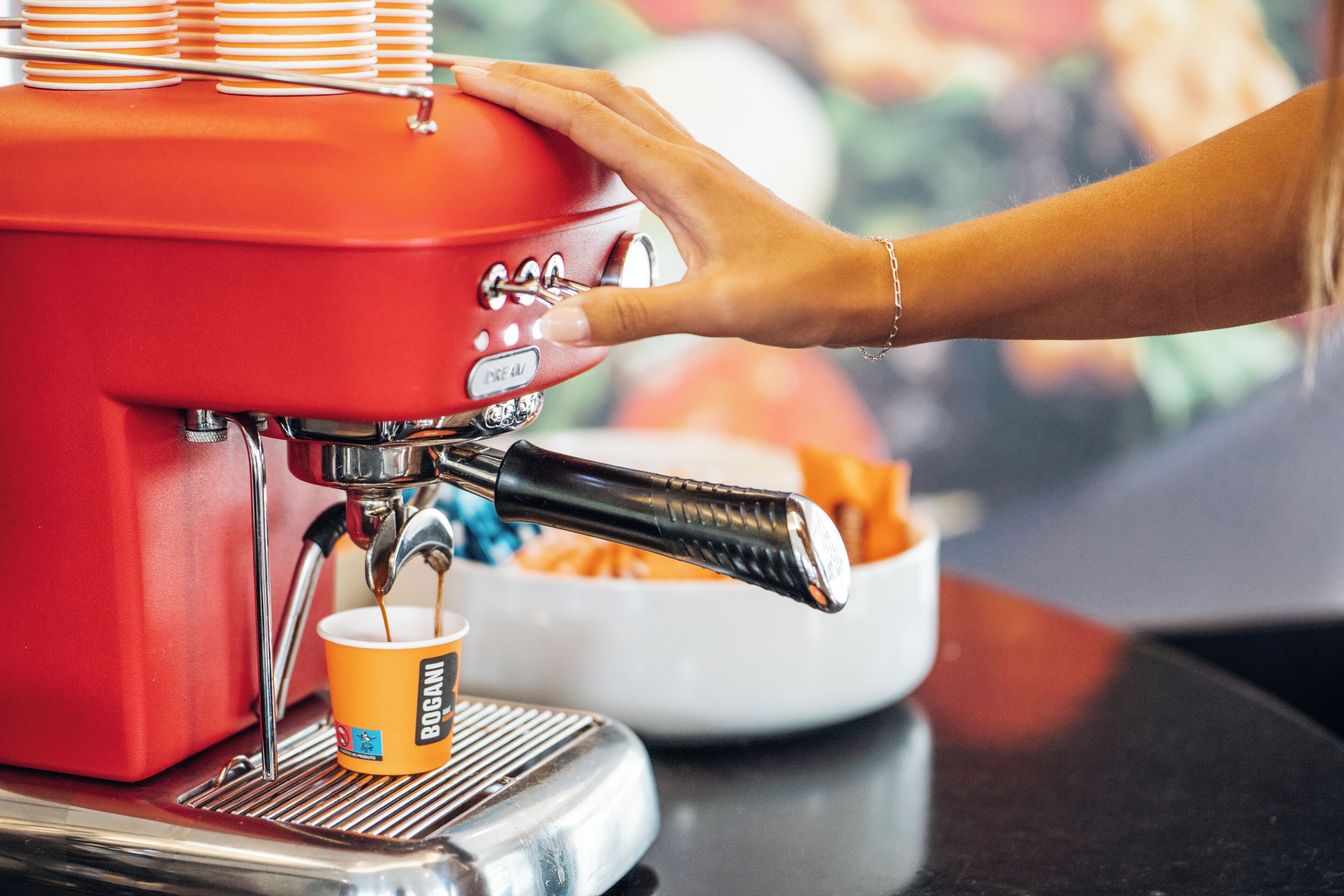 A hand operating a red espresso machine, with a cup labeled "BOGANI" and a bowl of coffee cups in the background.