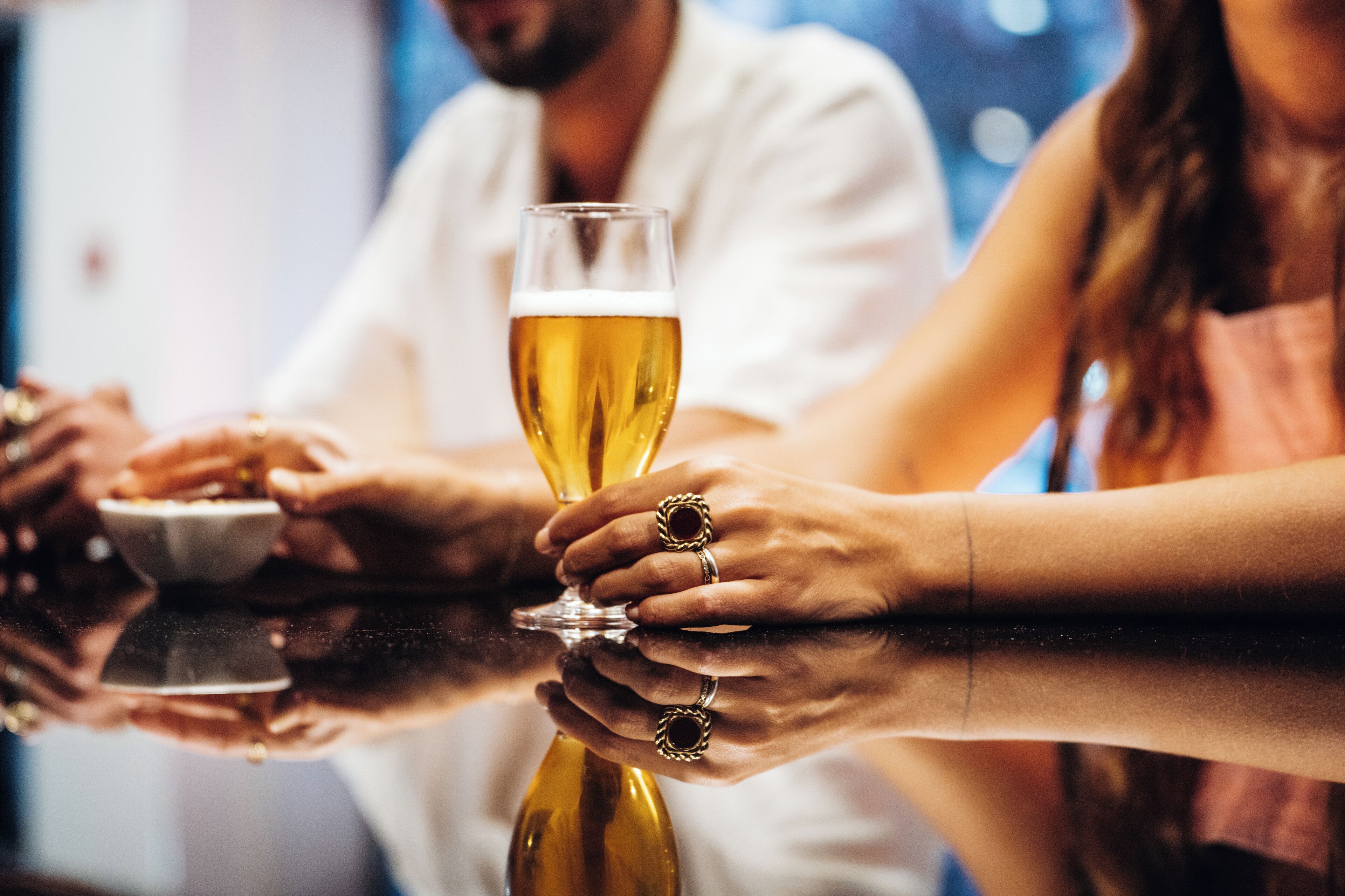 A close-up of hands holding a glass of beer, with a reflection on a polished surface.