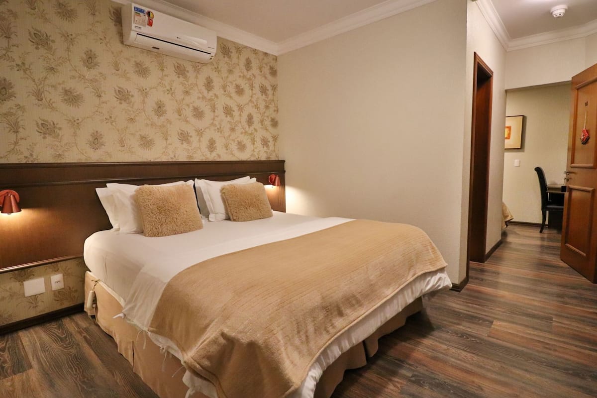 Une chambre d’hôtel avec un lit, un climatiseur, une tête de lit en bois et un couloir avec une porte.