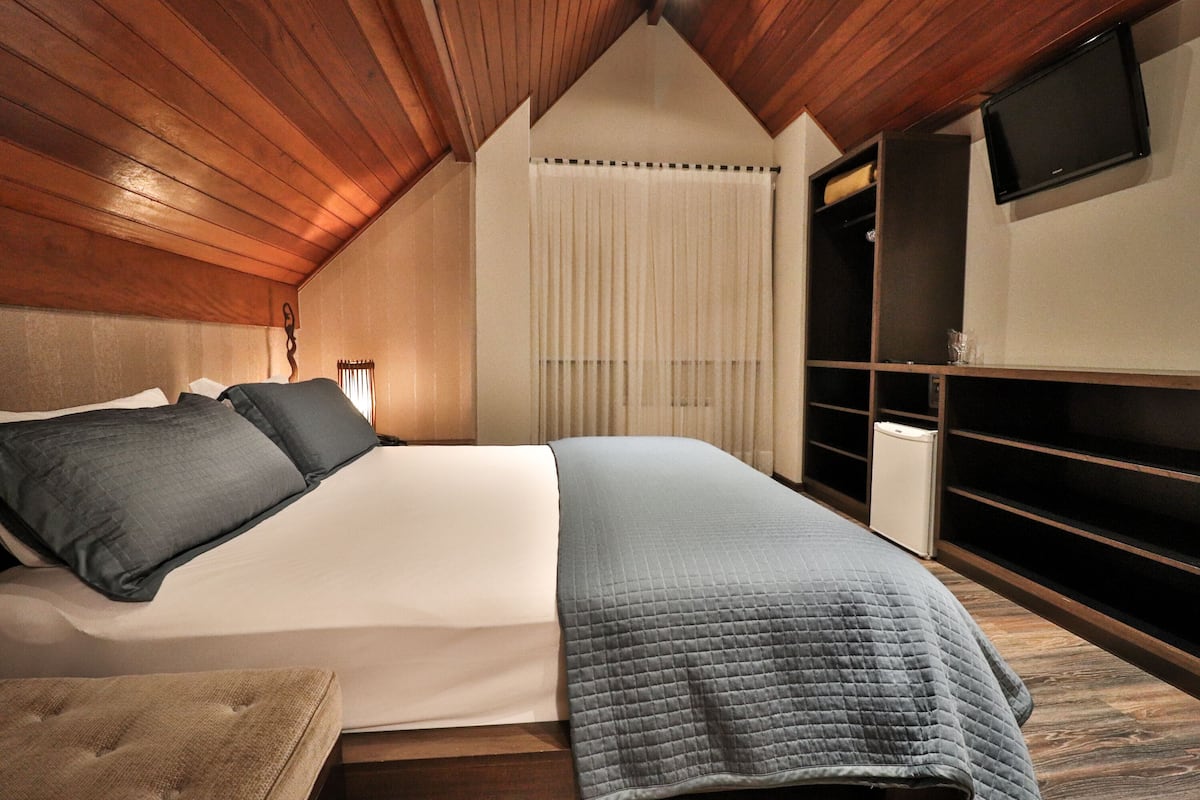 Une chambre avec un plafond en bois, un lit recouvert d’une couette blanche et bleue, une table de chevet, un téléviseur fixé au mur et une fenêtre avec des rideaux.