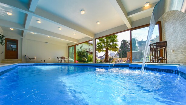 En swimmingpool med klart blåt vand, omgivet af et moderne interiør med store vinduer, en trappe og udsigt til omgivelserne.
