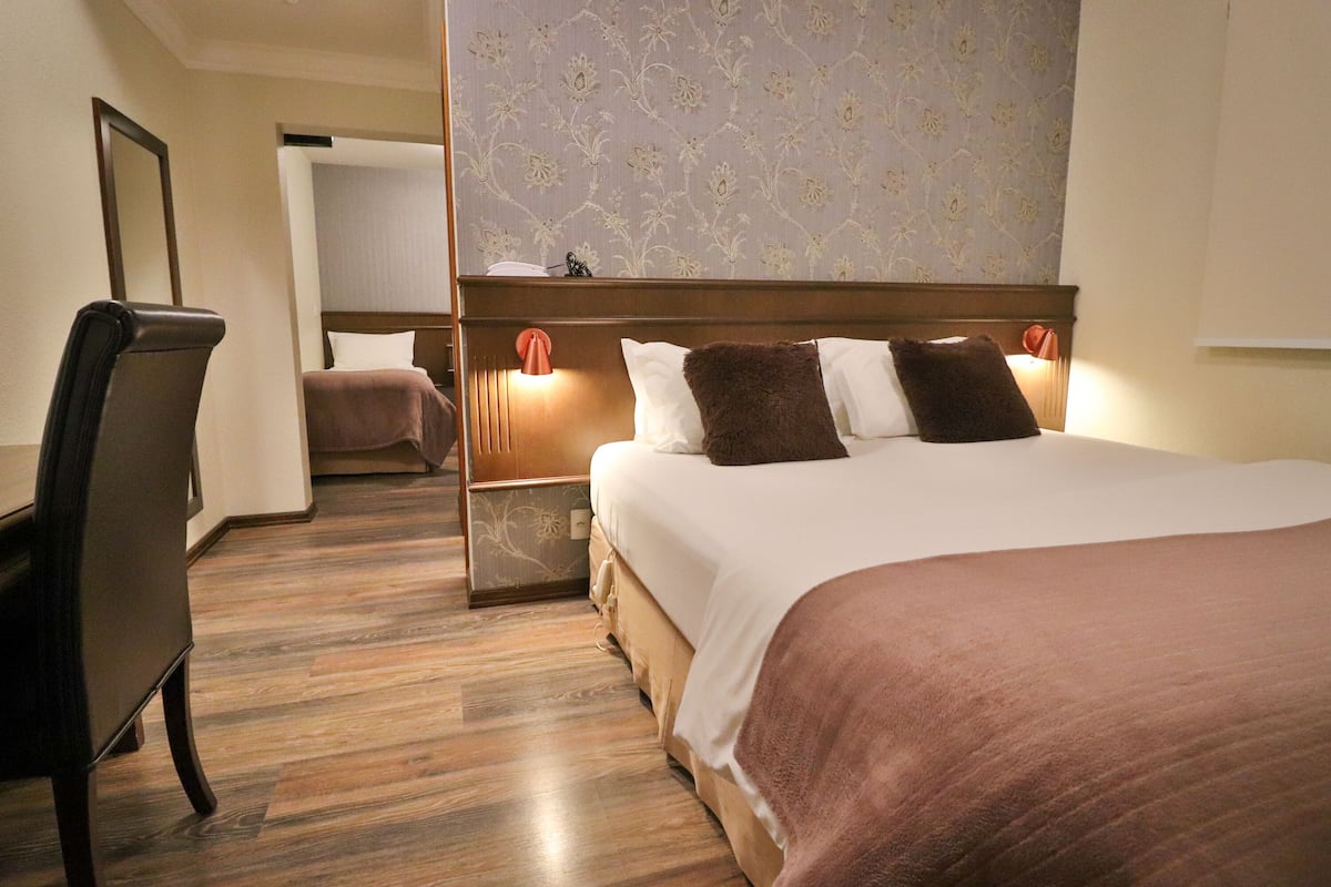 Une chambre d’hôtel avec un plancher en bois, un lit recouvert d’une couverture marron, une table de chevet avec une lampe et une chaise.