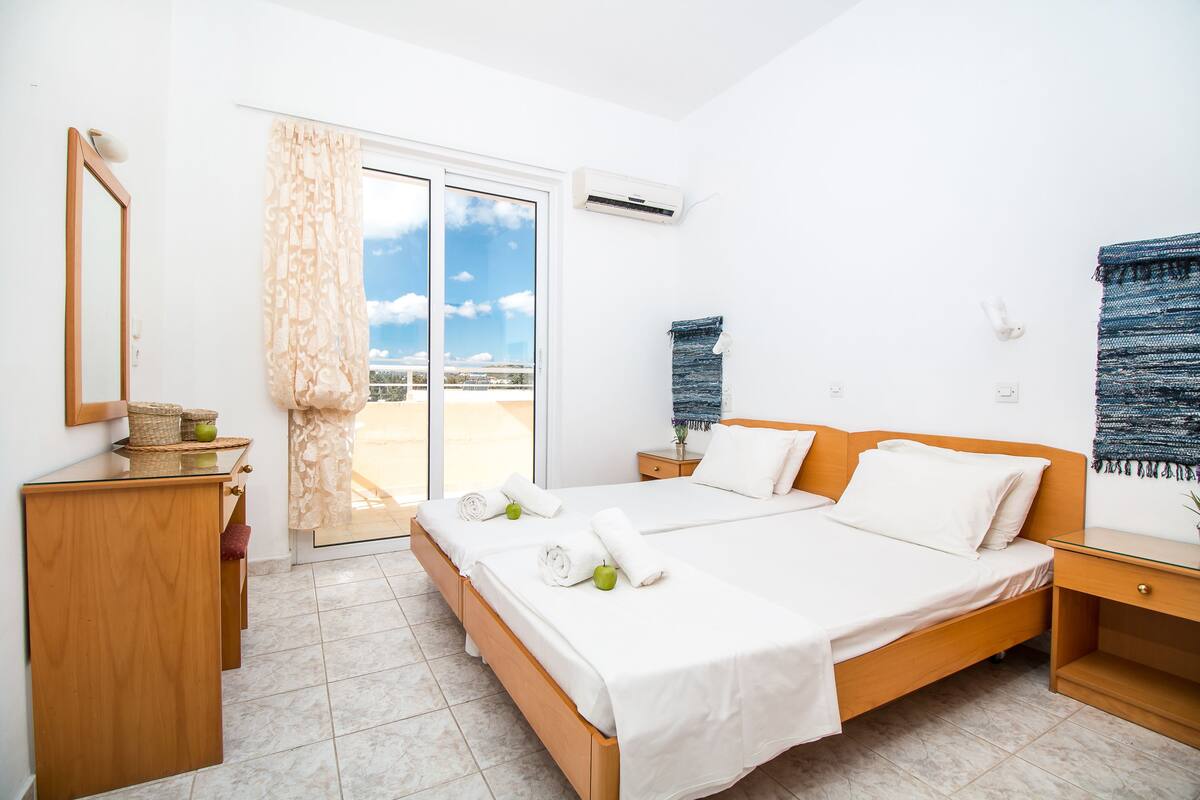 A hotel room with a large bed, a wooden nightstand, a small table, a mirror, and a sliding glass door leading to a balcony.