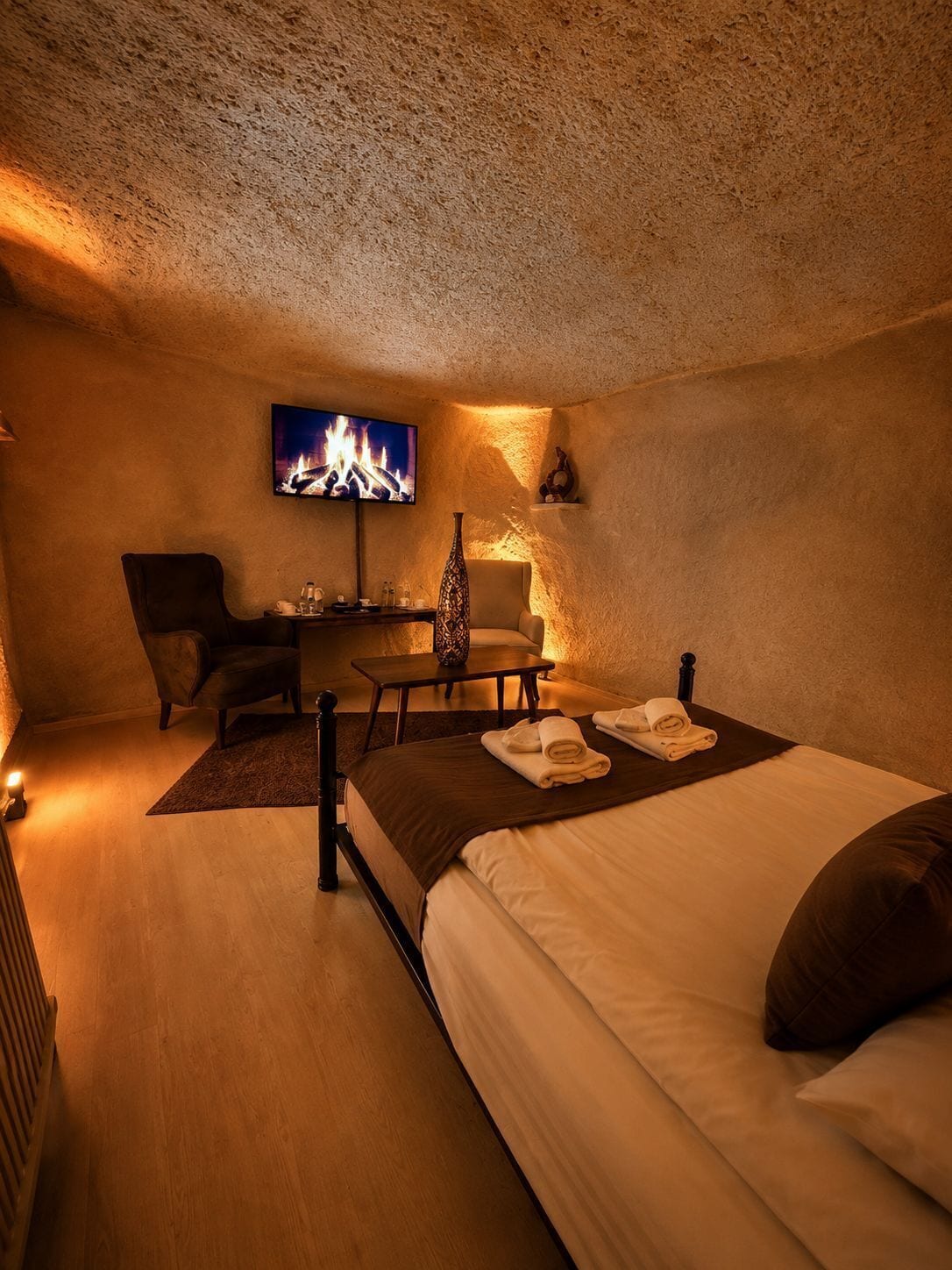 A cozy room with a bed, a chair, a small table, a TV displaying a fire, and a decorative vase.