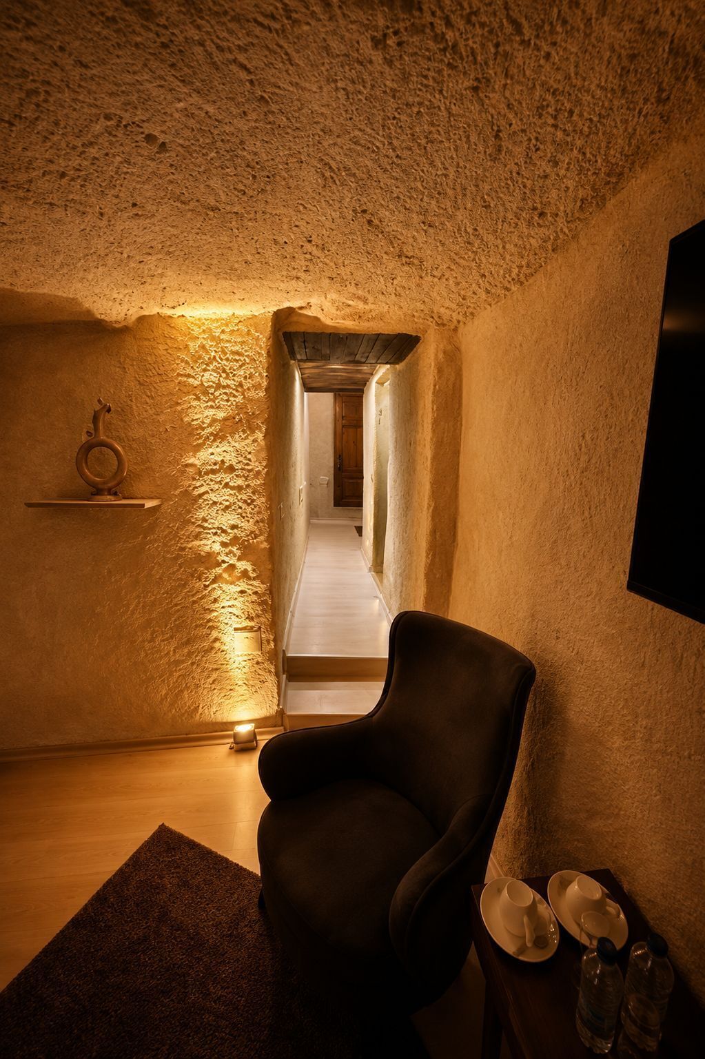 A cozy room with a chair, a small table with cups and a bottle, and a wooden shelf.