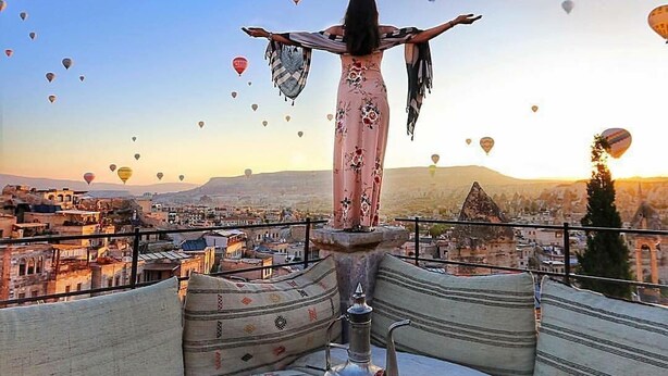 A person standing with arms outstretched on a balcony overlooking a cityscape with numerous hot air balloons in the sky. - Saklı Cave House (Avanos)