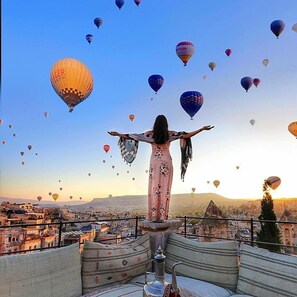 Una persona de pie, con los brazos extendidos, en un balcón que domina un paisaje urbano con numerosos globos aerostáticos en el cielo.