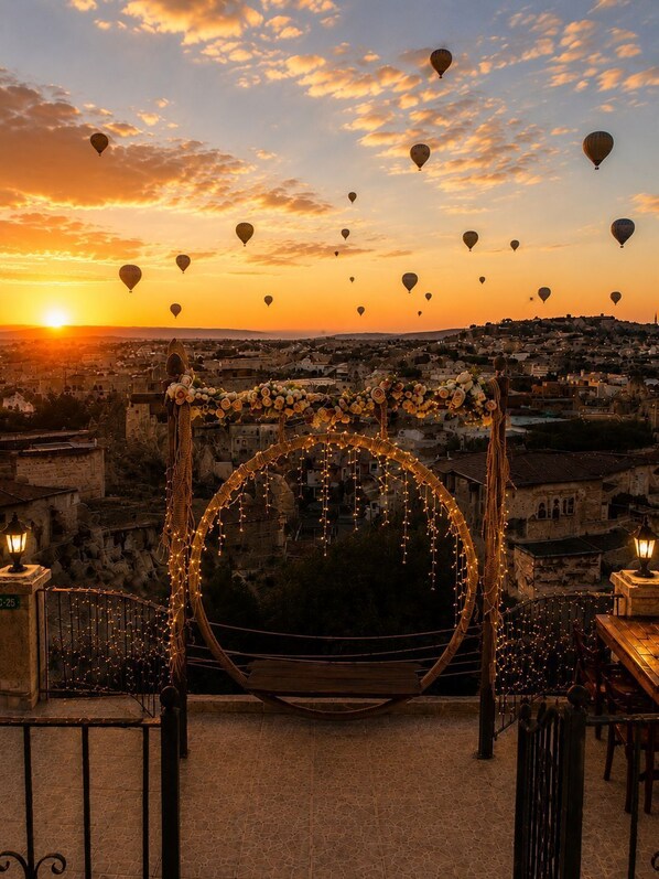 A balcony with a swing chair decorated with lights and flowers, overlooking a cityscape at sunset with hot air balloons in the sky. - Saklı Cave House (Avanos)