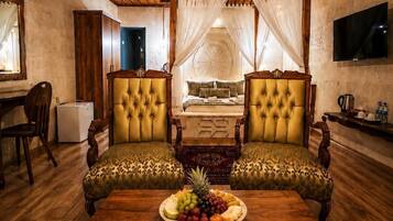 Una habitación con una cama, dos sillones, una mesa de madera con fruta y un televisor en la pared.