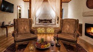 A room with a wooden ceiling, a bed with a canopy, two leather armchairs, a coffee table with fruit and wine glasses, and a fireplace. - Saklı Cave House (Avanos)
