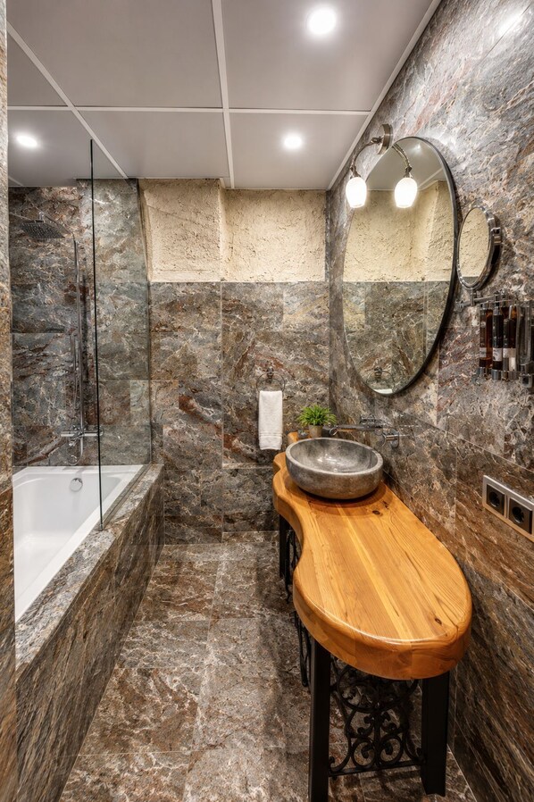 A bathroom with a bathtub, a wooden vanity, a round mirror, and a stone wall.