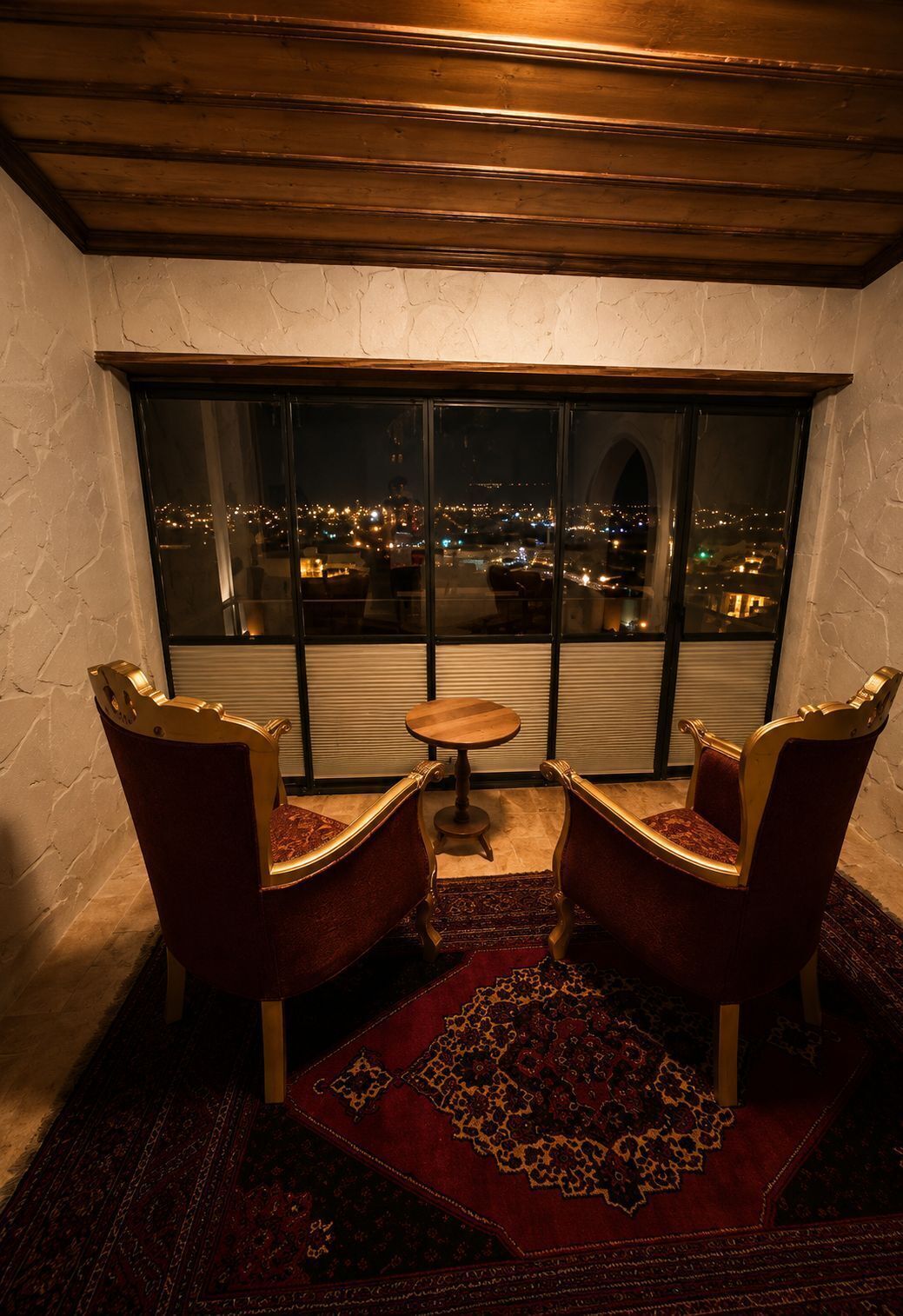 Una habitación con dos sillones de terciopelo rojo, una pequeña mesa de madera y un ventanal grande con vista a la ciudad de noche.
