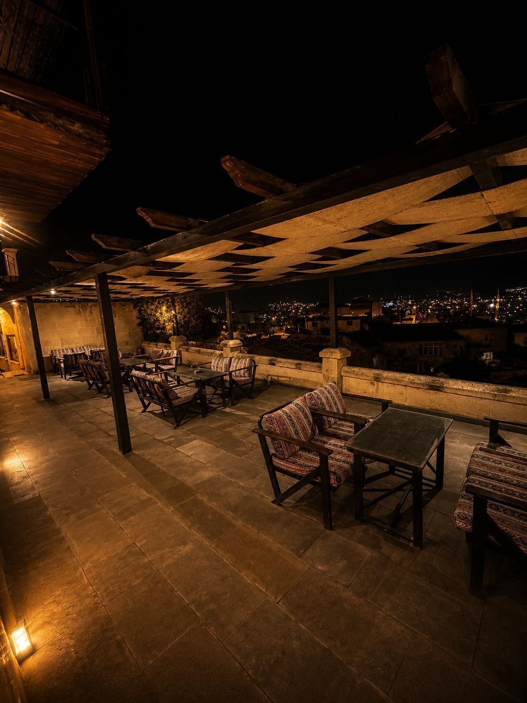 Un patio al aire libre con suelo de piedra, mobiliario de madera y vistas a un paisaje urbano nocturno.