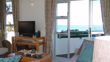 Una sala de estar con televisor sobre un mueble de madera, un sofá con cojines estampados y vista al mar a través de amplios ventanales.