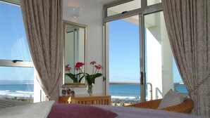Ein Schlafzimmer mit Bett, Kommode, Blumen in einer Vase und einem großen Fenster mit Vorhängen.