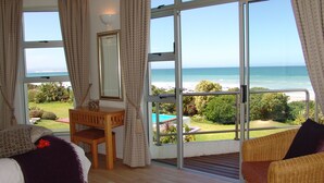 Ein Zimmer mit großem Fenster mit Blick auf einen Strand, einem Holztisch und einem Korbstuhl.