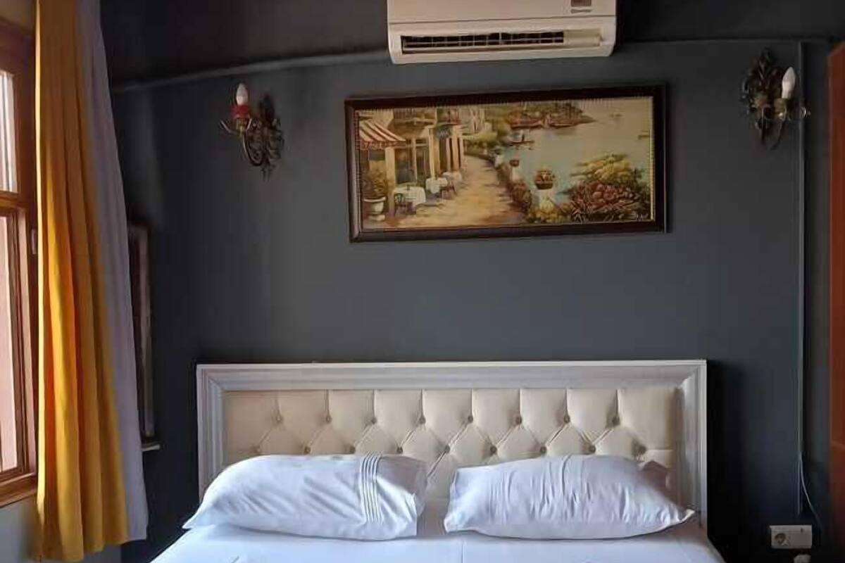 A hotel room with a large bed, white linens, a tufted headboard, an air conditioning unit, and a painting on the wall.