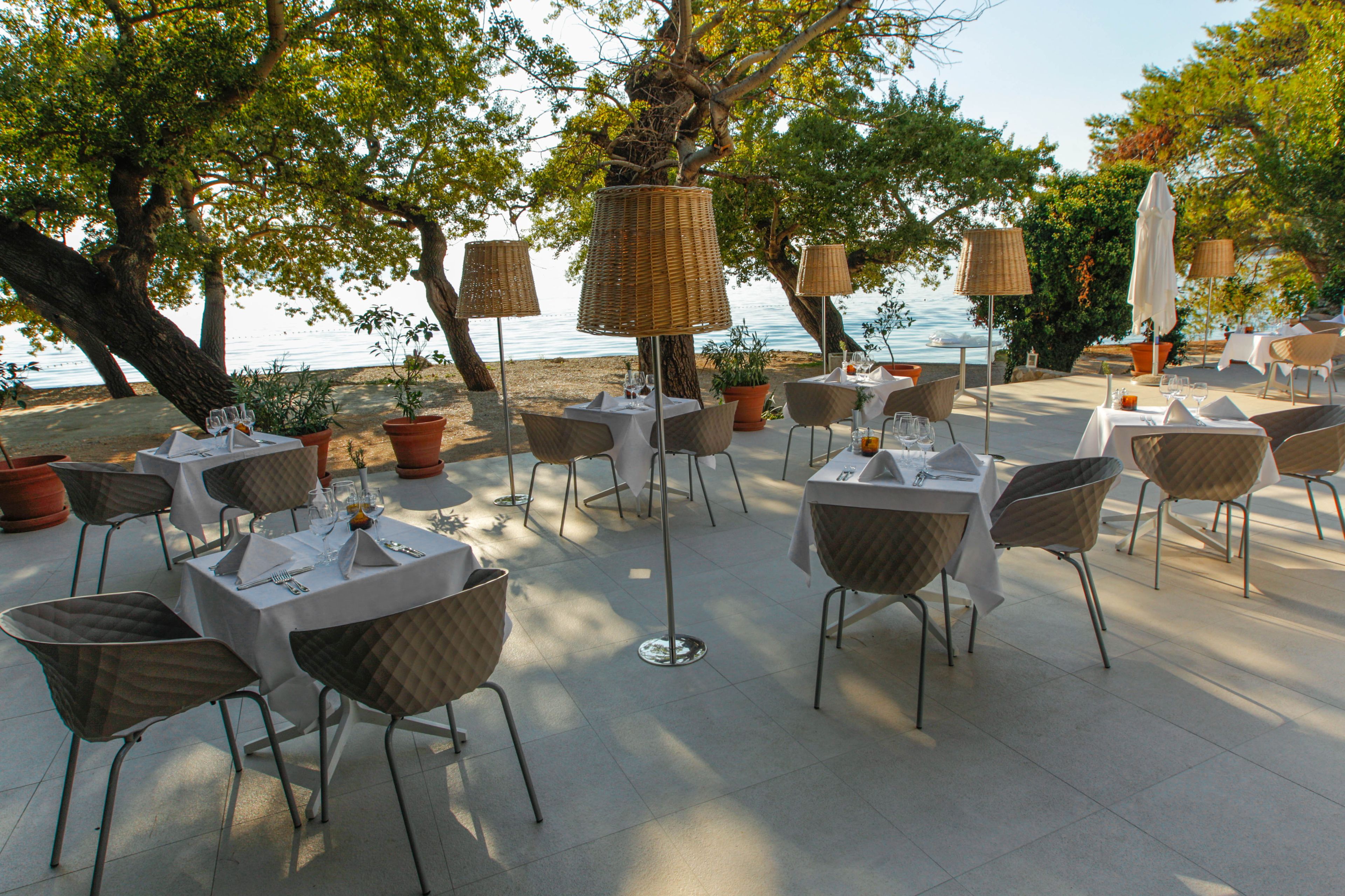 An outdoor dining area with tables and chairs, surrounded by trees and overlooking a body of water.