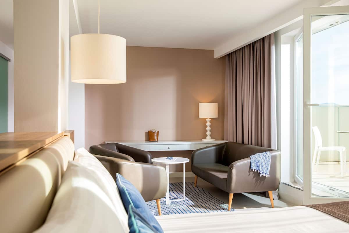 A modern hotel room with a sofa, two armchairs, a small table, a lamp, and a view of the balcony.