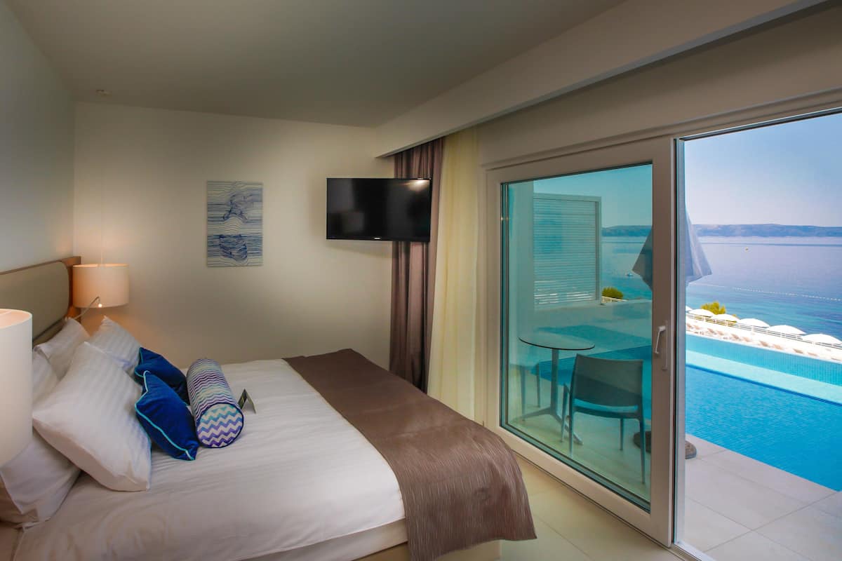 A hotel room with a bed, a TV mounted on the wall, a balcony with a view of the sea, and a small table with chairs.