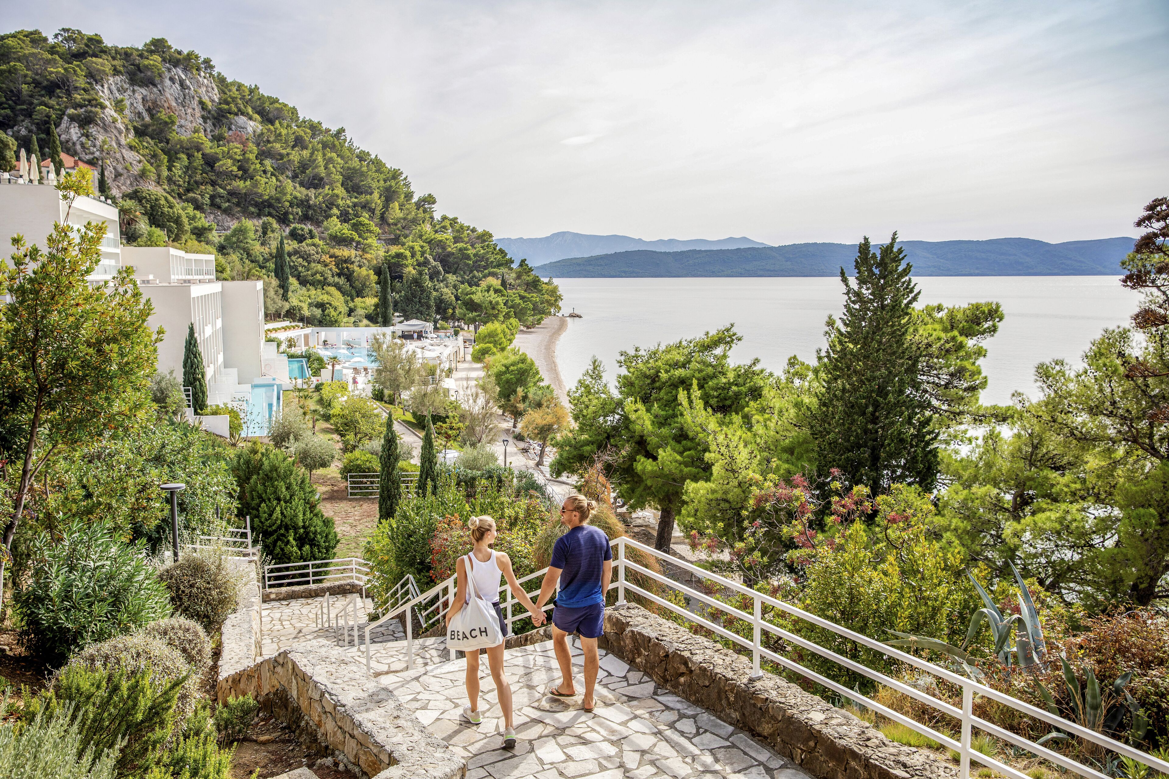Un couple se promène sur un escalier en pierre qui surplombe une plage, offrant une vue sur un lac et des montagnes.