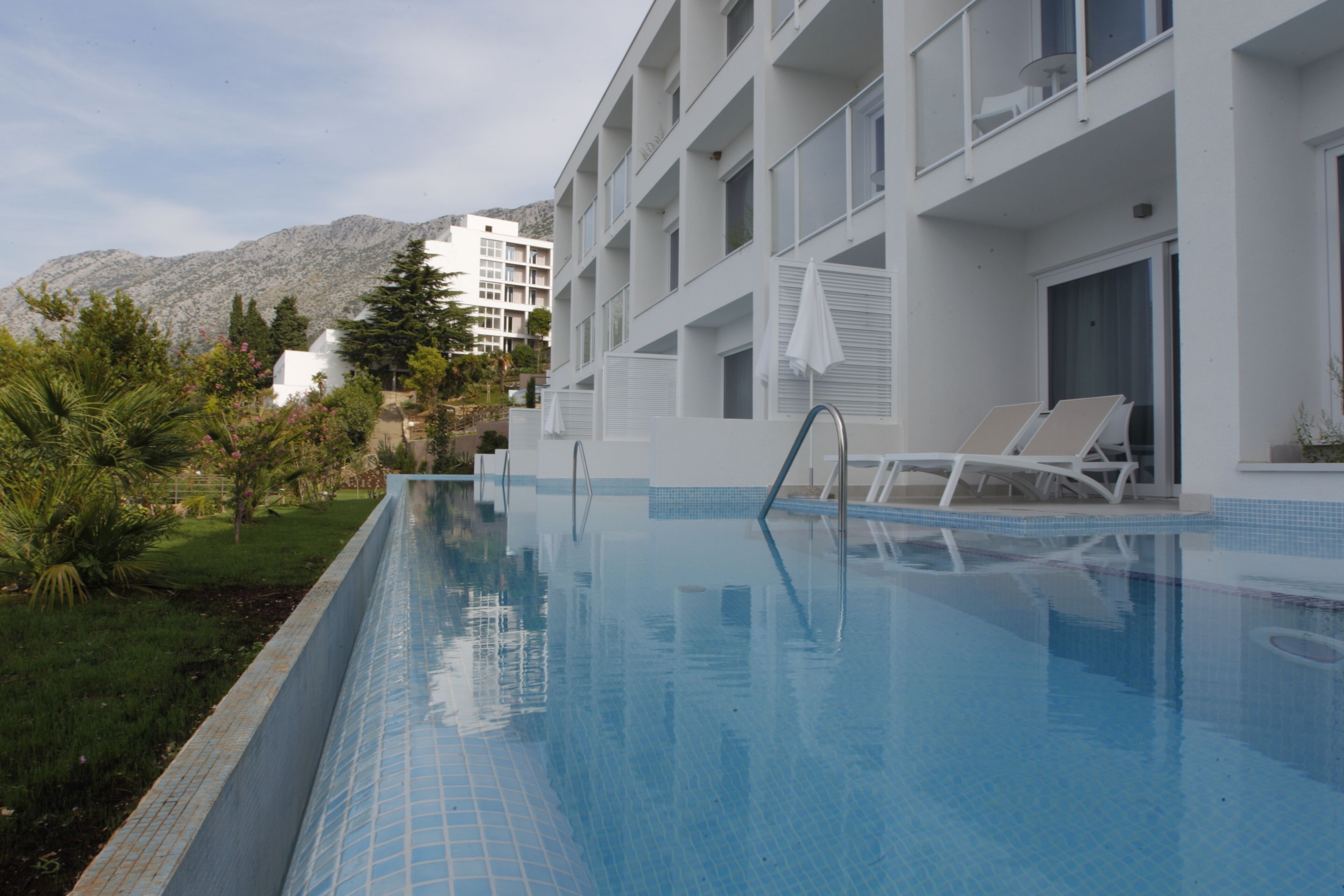 A swimming pool with lounge chairs, a white building with balconies, and a mountainous backdrop.
