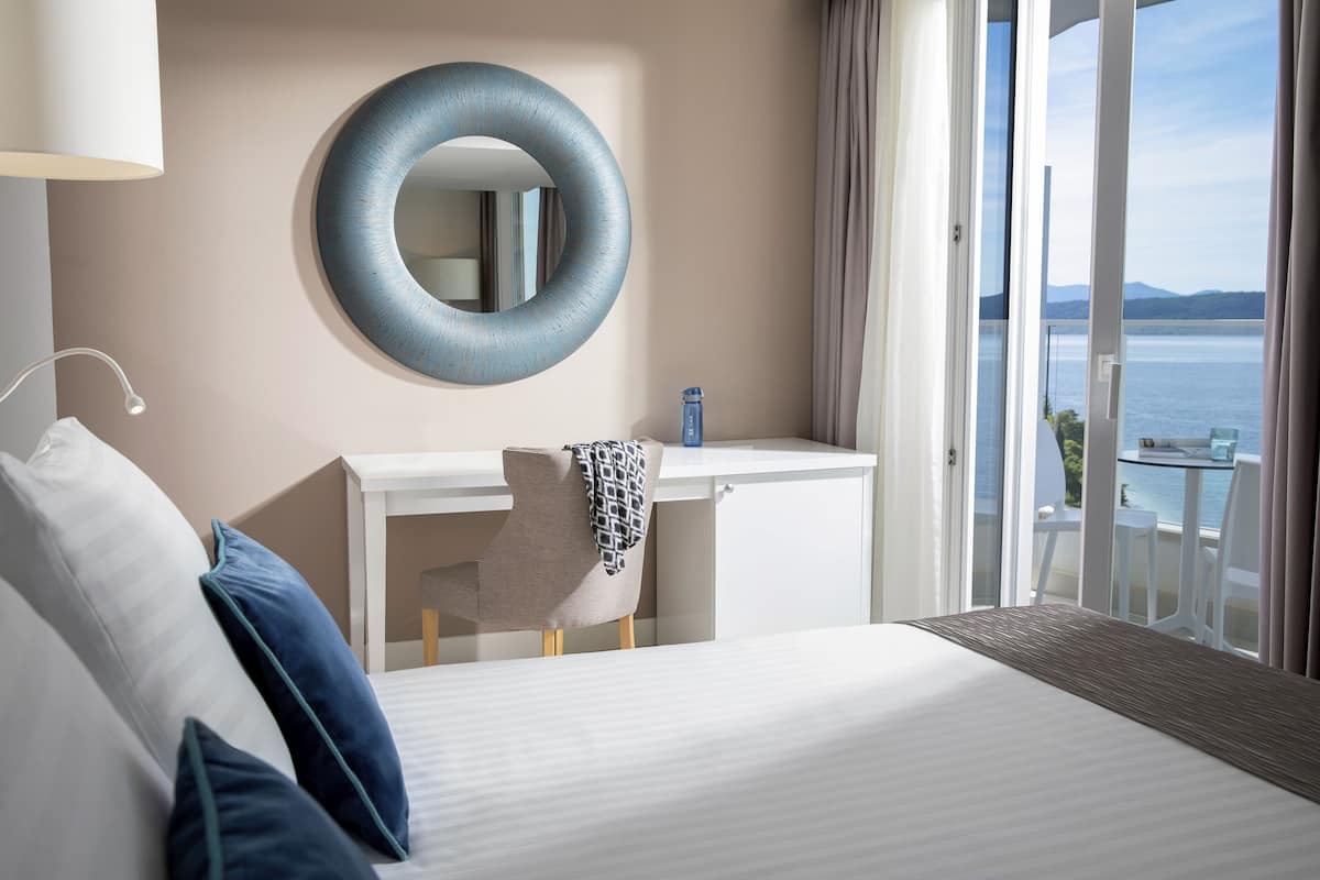 A hotel room with a bed, a desk, a chair, a round mirror, and a view of the sea.