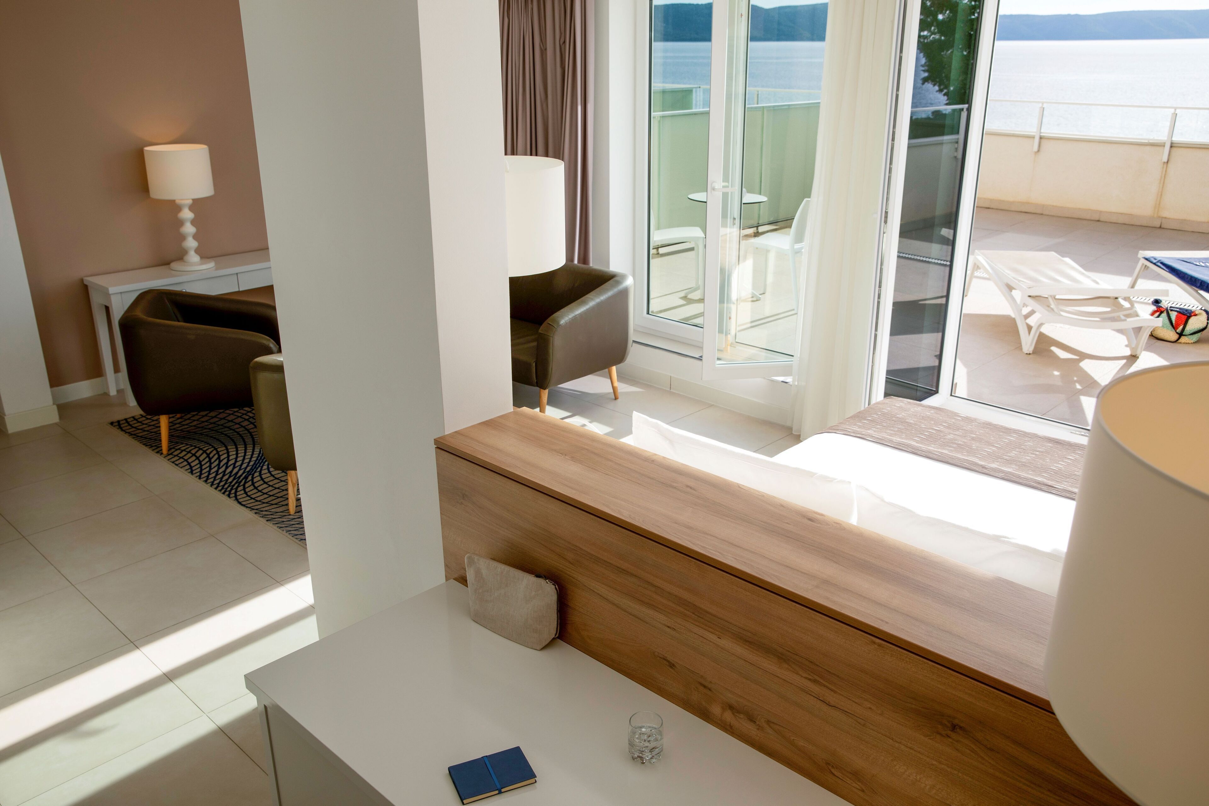 A modern hotel room with a wooden desk, a chair, a sofa, and a view of the sea through large glass doors.