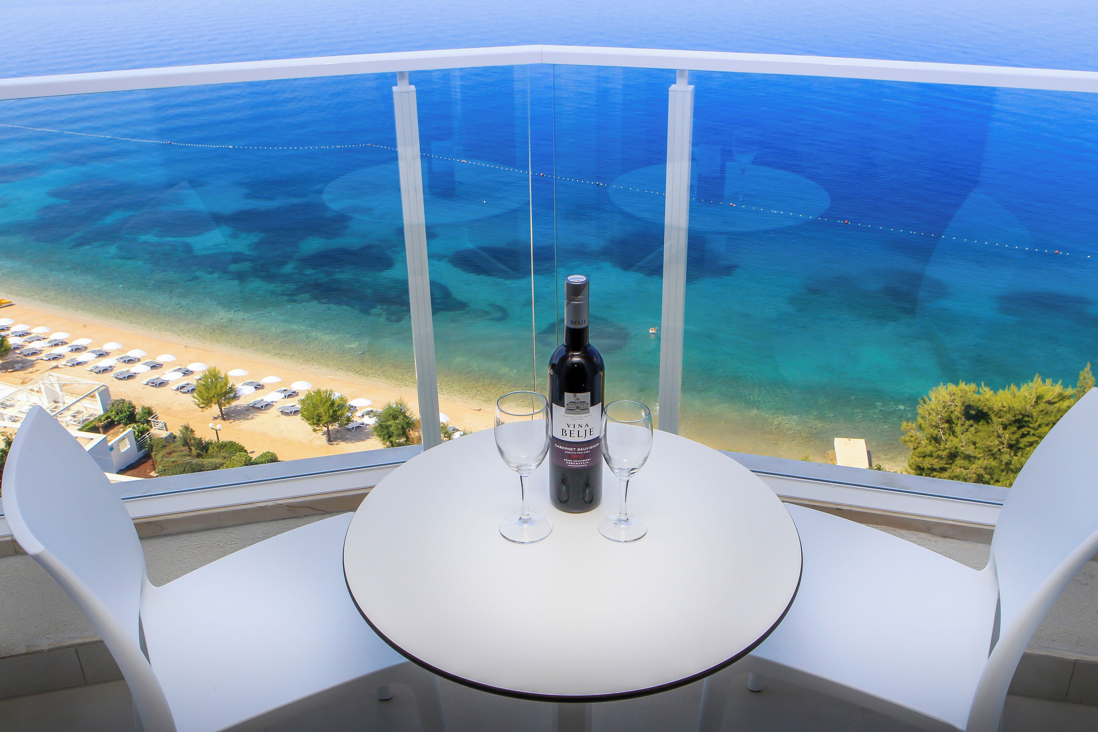 Een balkon met een tafel en twee stoelen, een fles wijn, en uitzicht op een strand en zee.