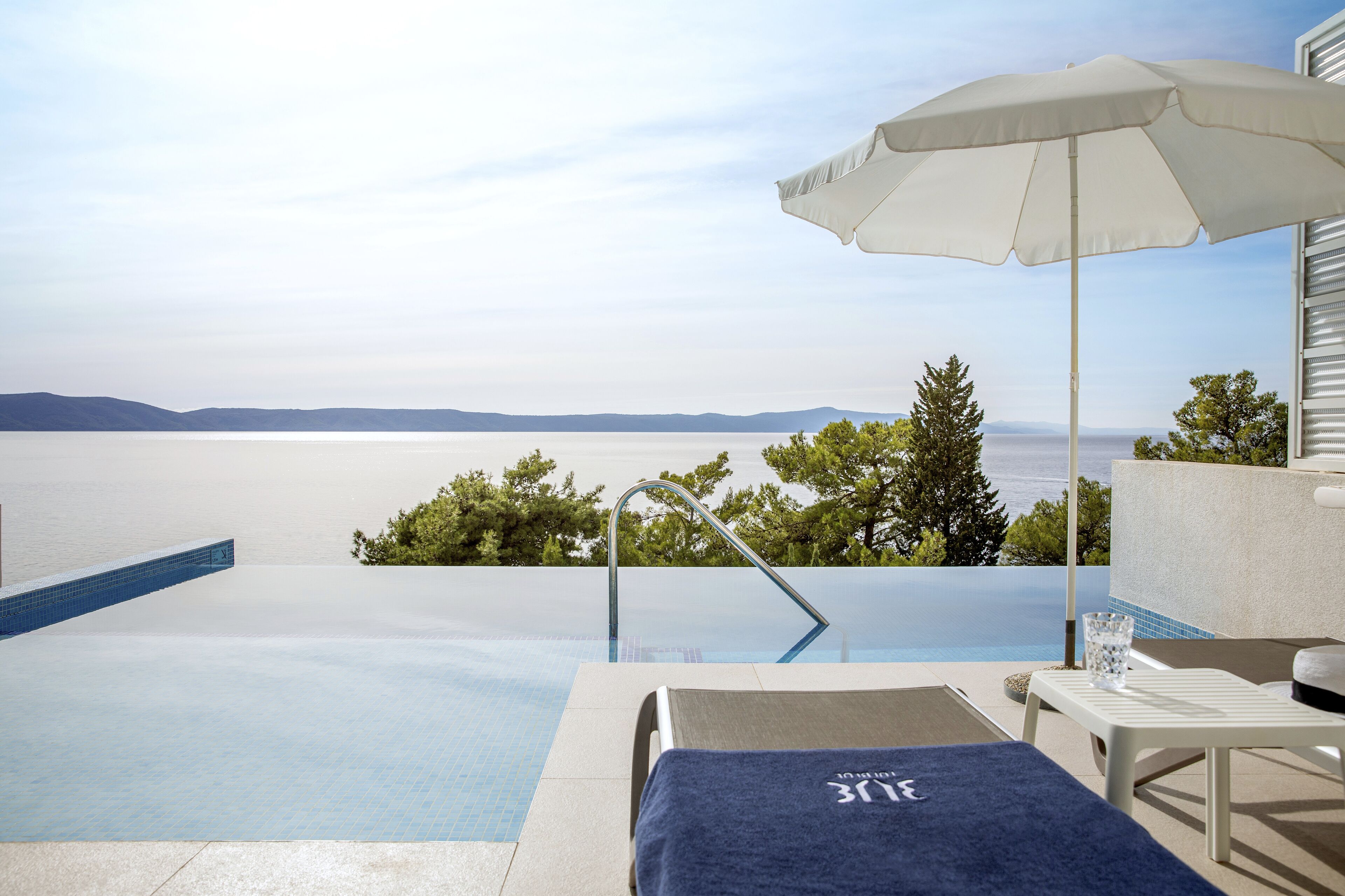 A poolside area with a view of the sea, a lounge chair, a small table, and an umbrella.