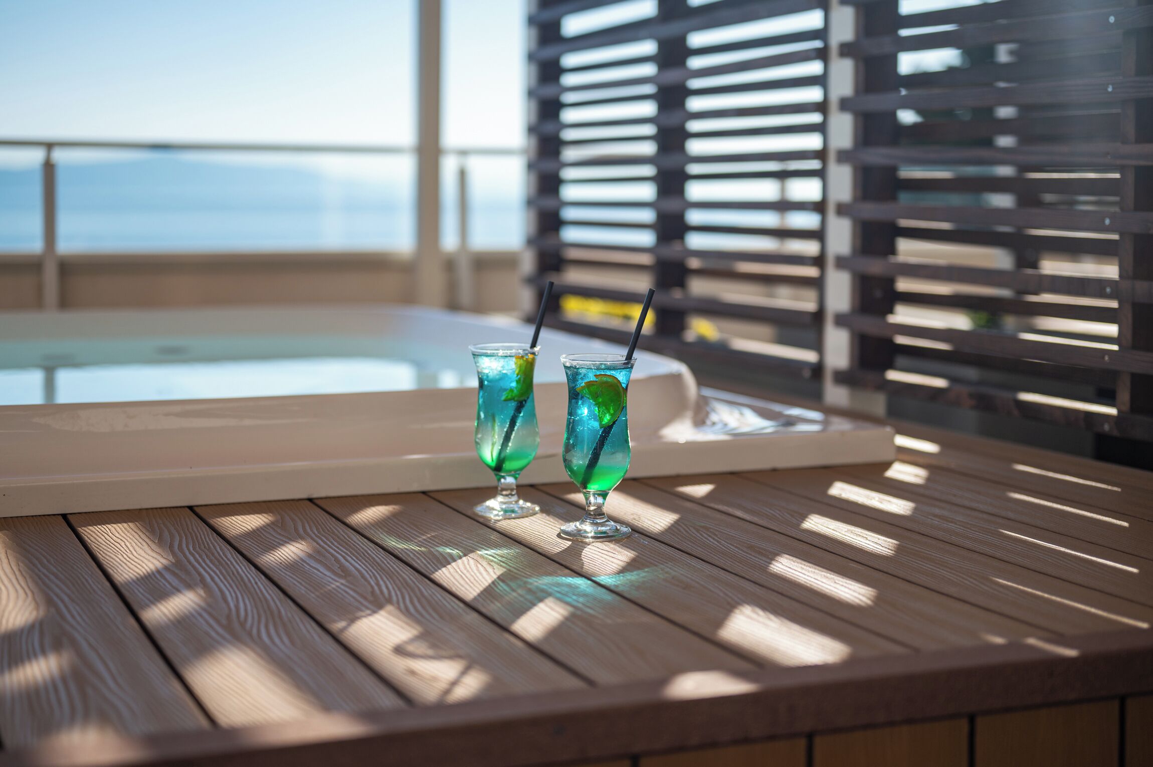 Two glasses of blue cocktail on a wooden table with a view of a pool and a building in the background.