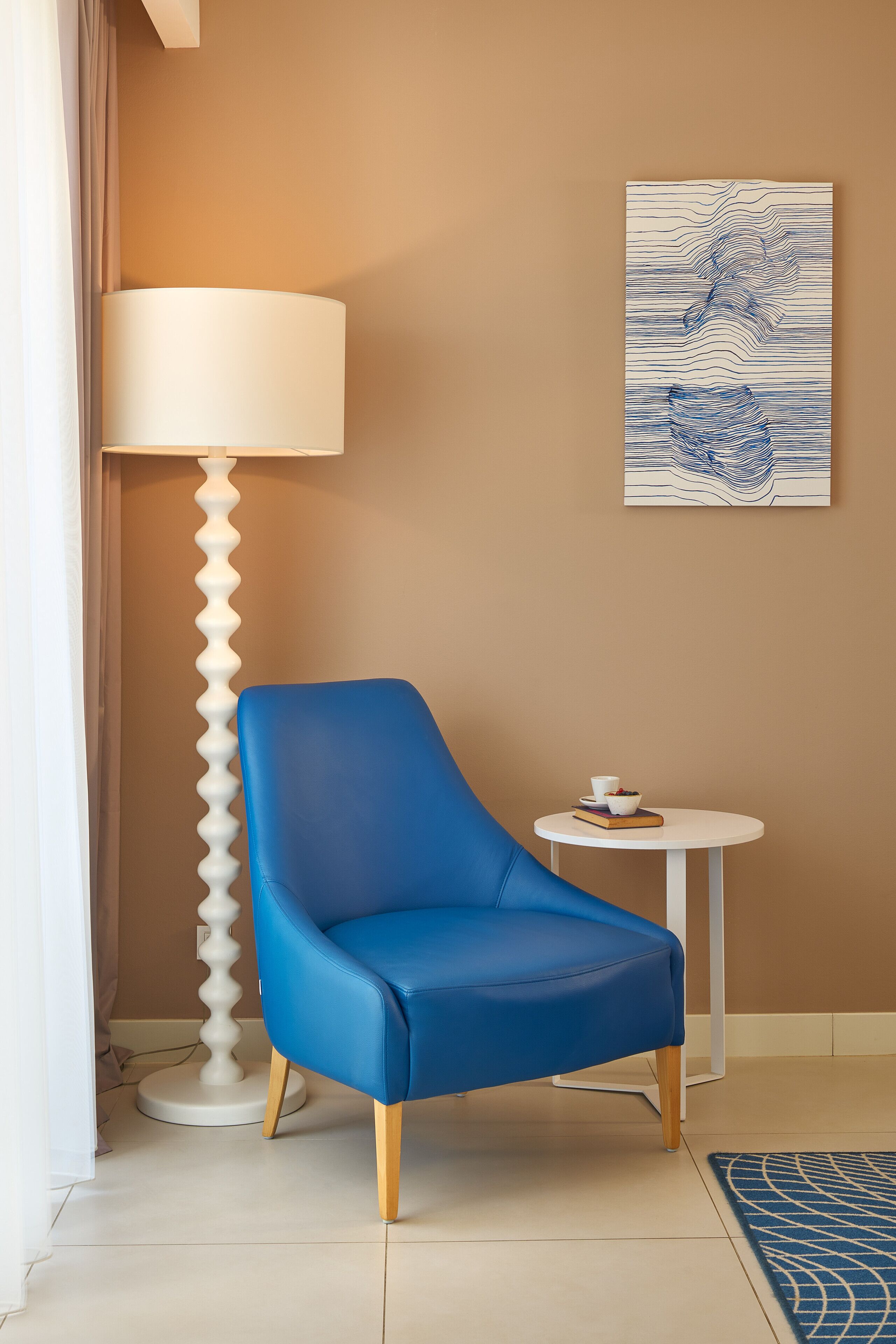 A blue chair, a white lamp, a small round table with a cup and book, and a blue patterned rug.