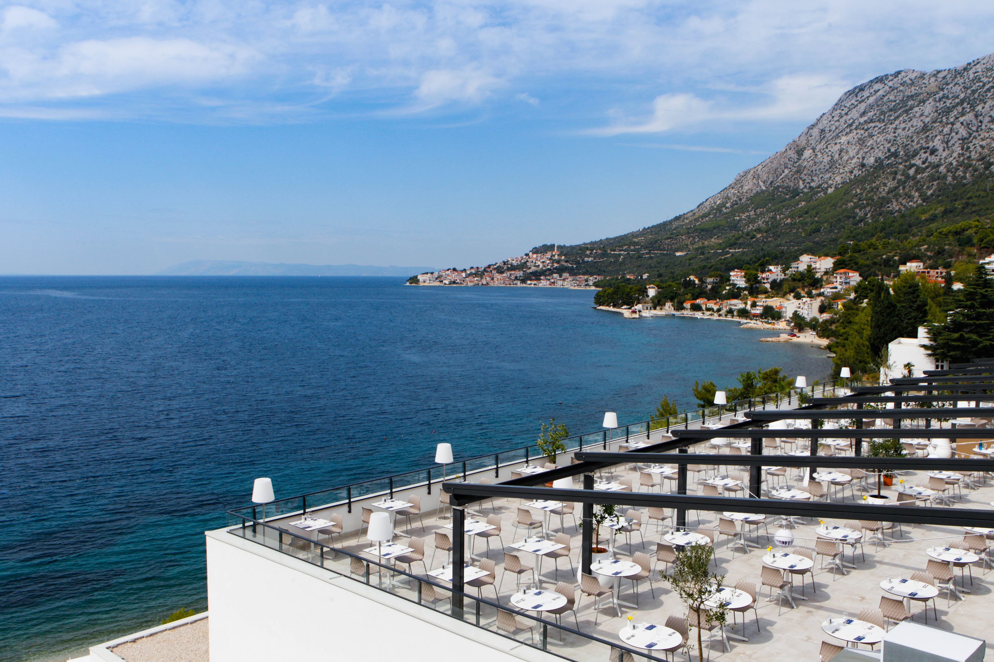 A seaside terrace with outdoor seating and a view of the sea and a mountainous backdrop.