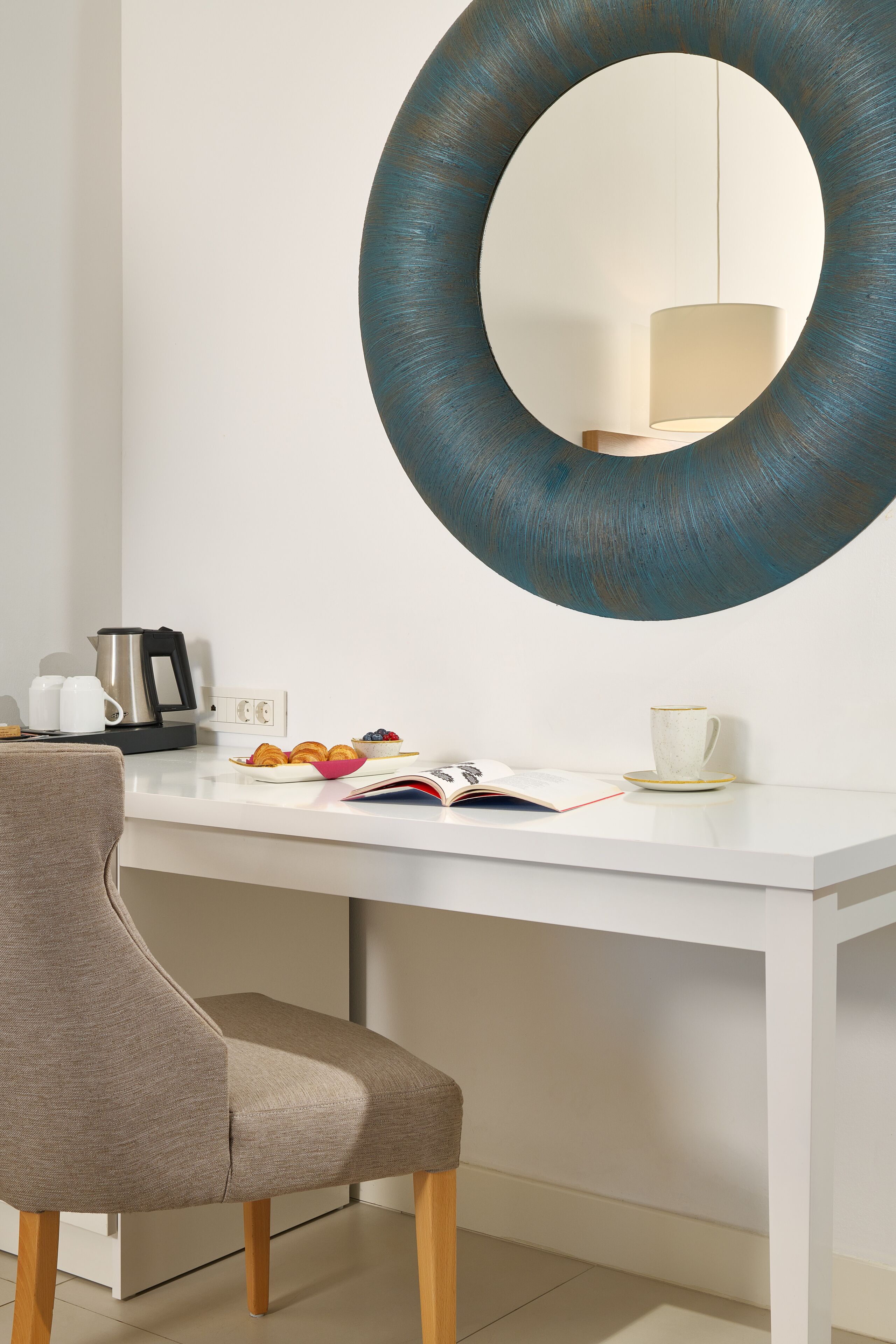 A modern room with a white desk, a large mirror, a chair, and a coffee maker.