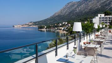 A seaside terrace with outdoor seating, overlooking a calm sea and a mountainous backdrop.