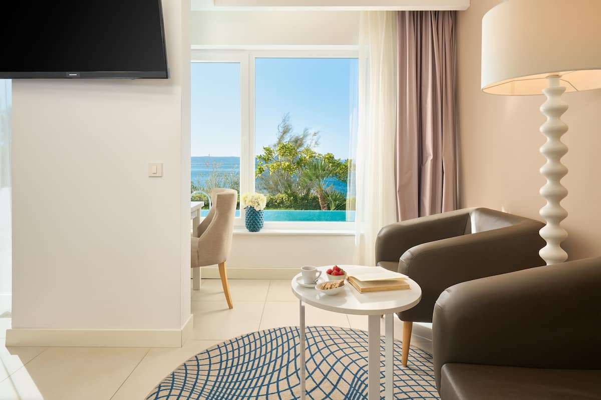 A modern living room with a flat-screen TV, a round coffee table with a book and a cup, two sofas, a lamp, and a view of the sea through large windows.