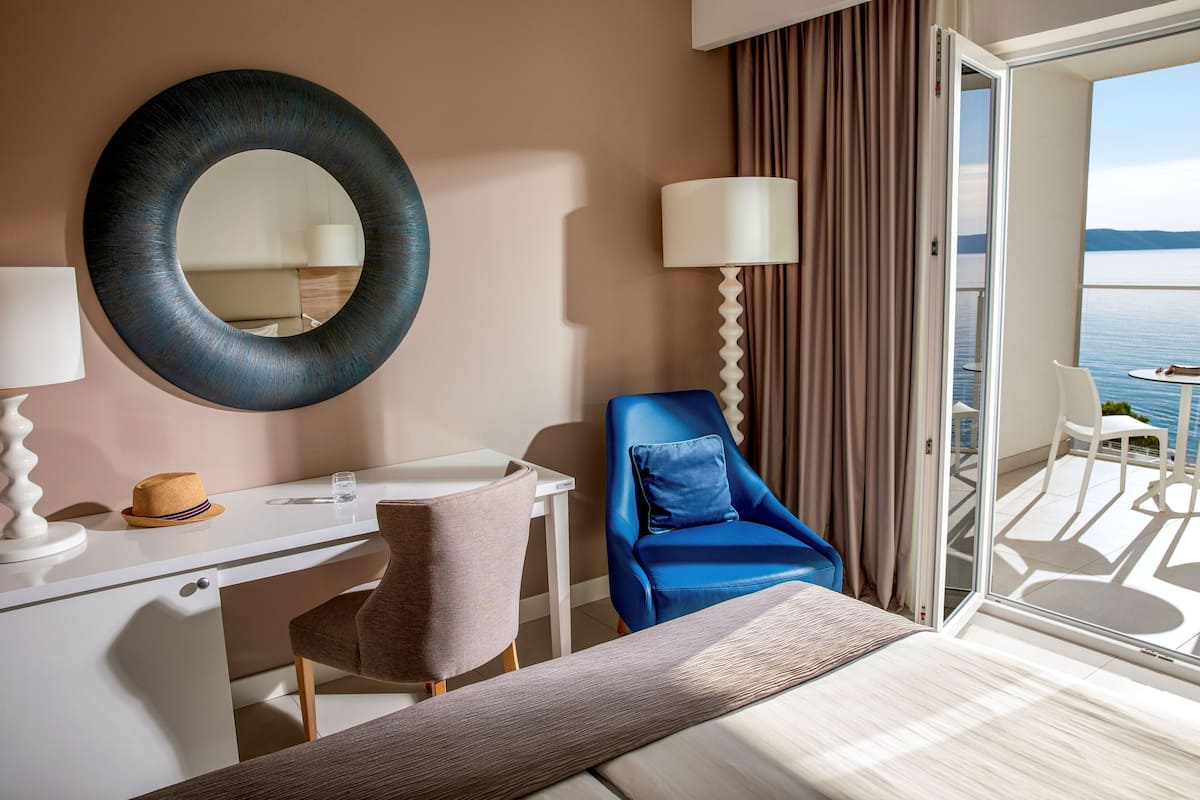 A hotel room with a bed, a desk, a chair, a large mirror, a lamp, and a view of the sea through a balcony.