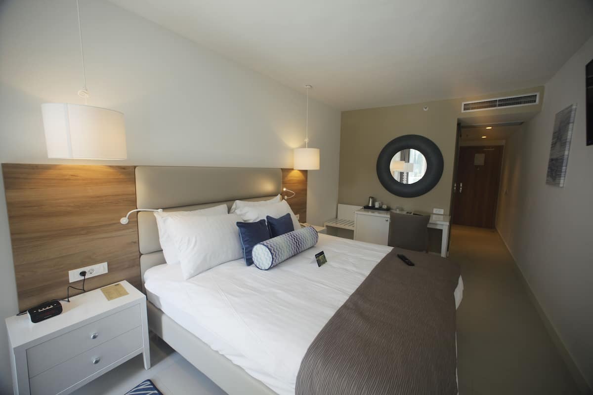 A modern hotel room with a large bed, bedside tables, a round mirror, and a wooden headboard.
