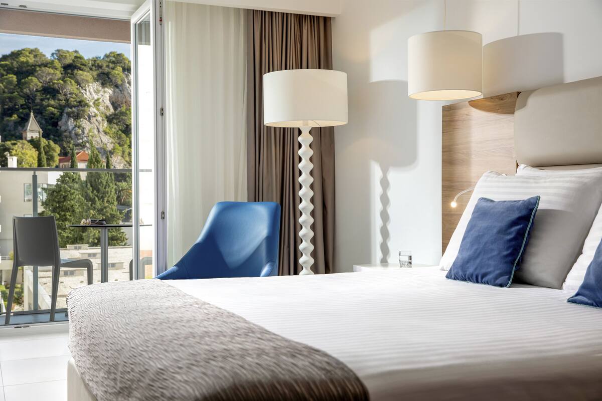A hotel room with a large bed, a chair, a table, and a view of a mountainous landscape.