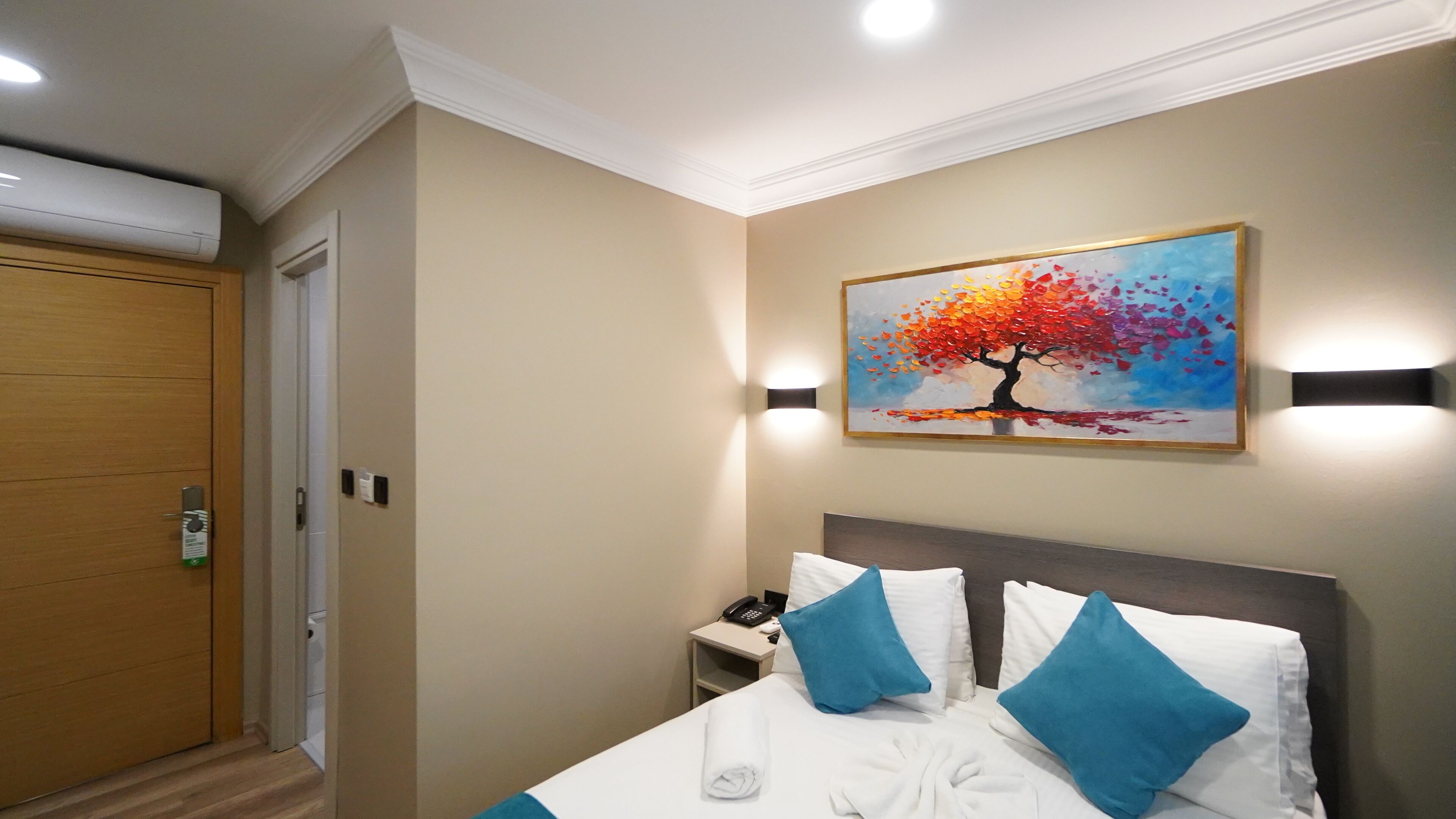A hotel room with a bed, a painting of a tree, a bedside table with a phone, and a door.