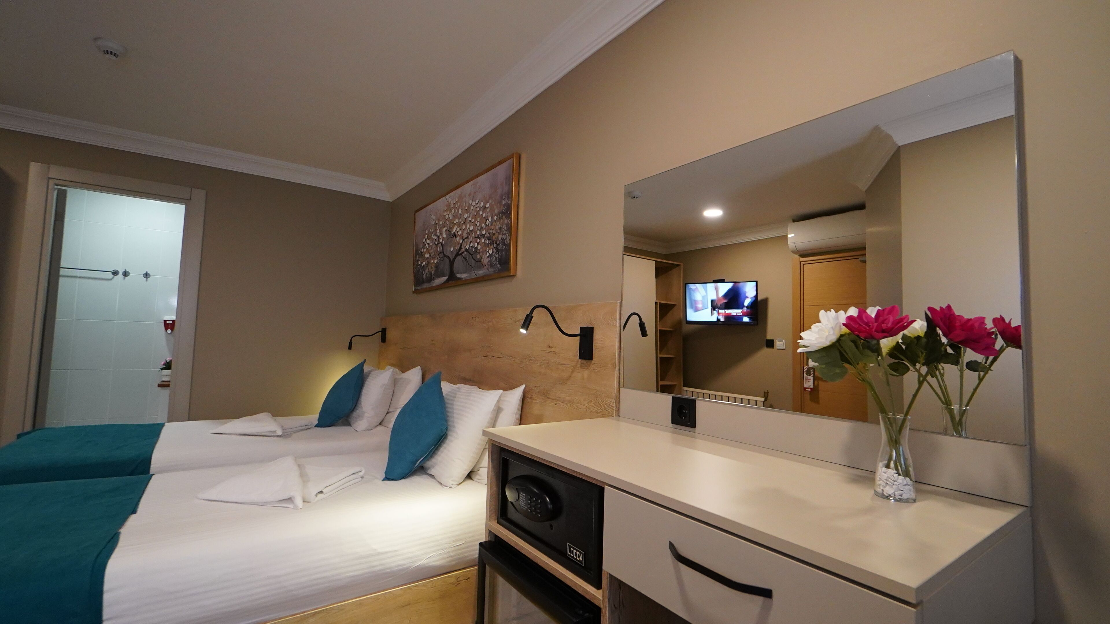 A hotel room with a bed, a desk, a mirror, a TV, and a flower vase.