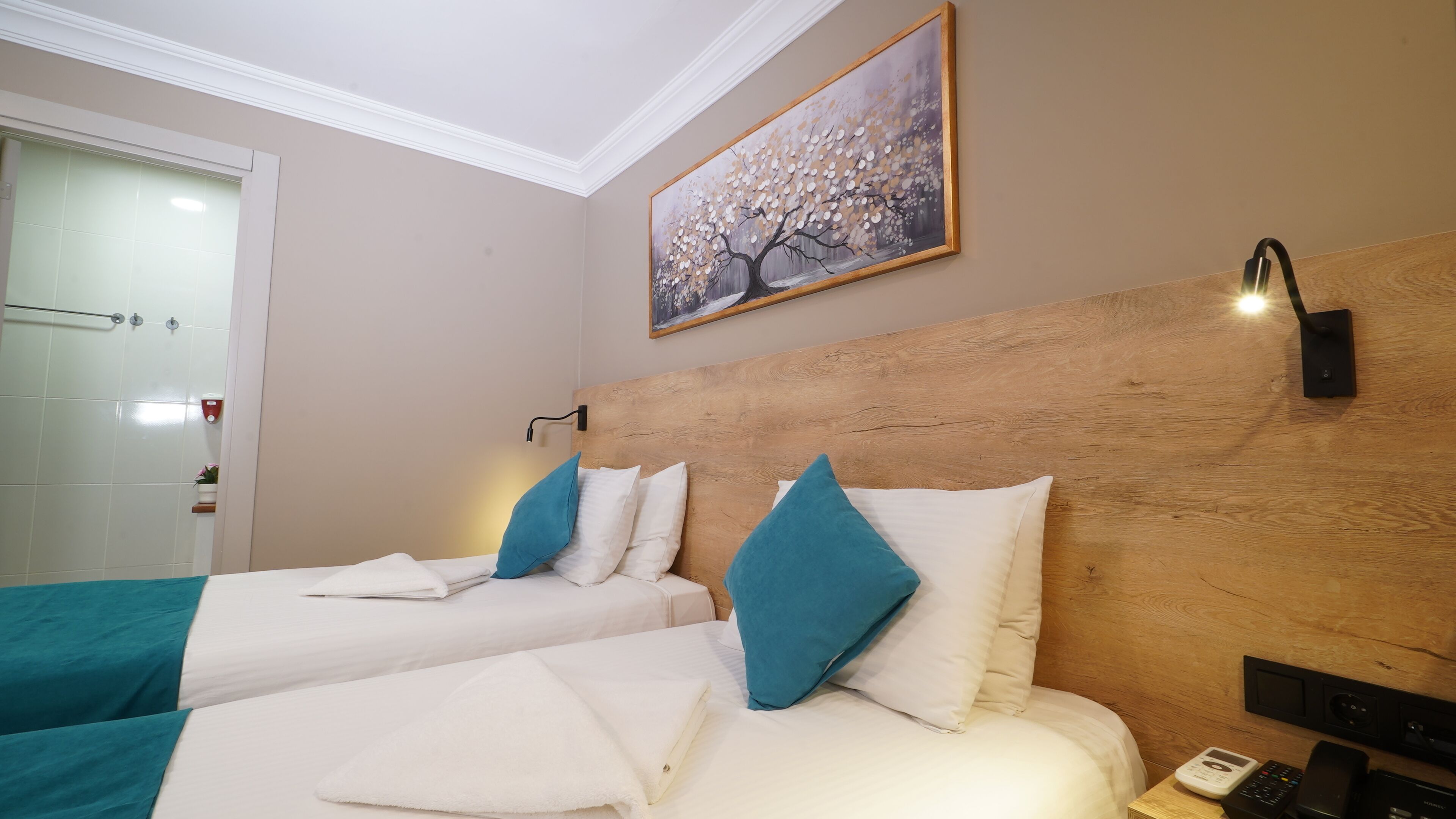 A hotel room with two beds, a wooden headboard, a painting above the beds, a nightstand with a phone, and a bathroom with a glass door.