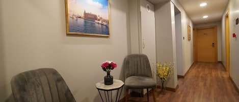 A hotel hallway with a wooden floor, two armchairs, a small table with a vase of flowers, and a painting of a cityscape on the wall.