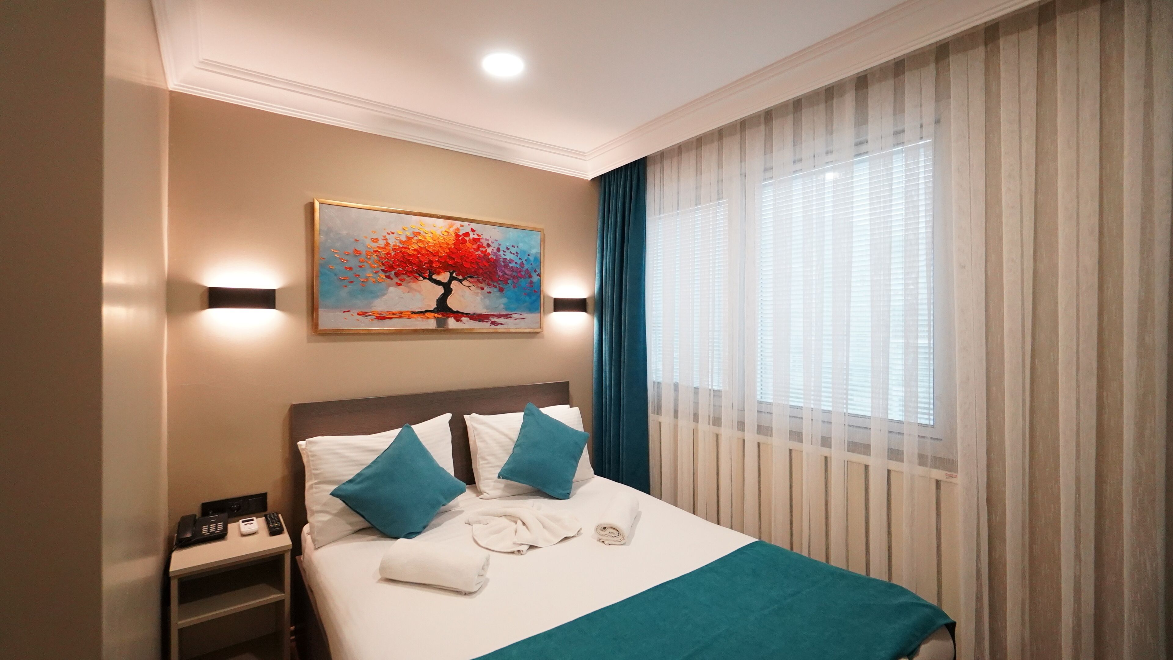 A hotel room with a bed, a painting of a tree, a nightstand with a phone, and a window with curtains.