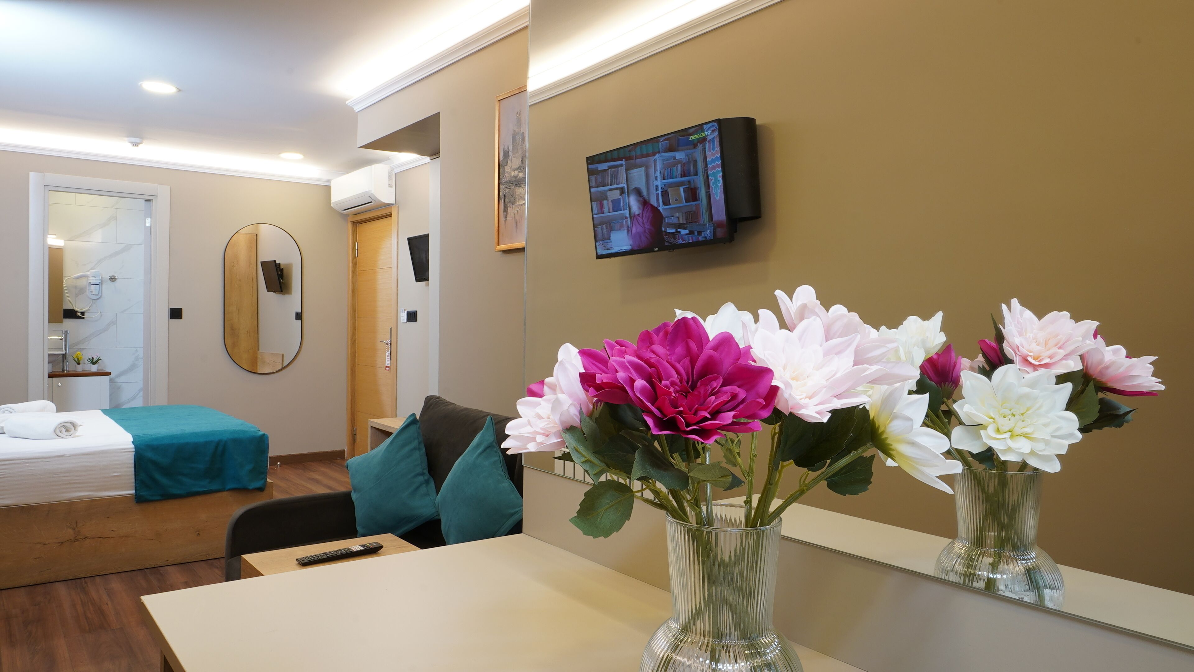 Ein Hotelzimmer mit Bett, Wandfernseher, Spiegel und Blumenvase.