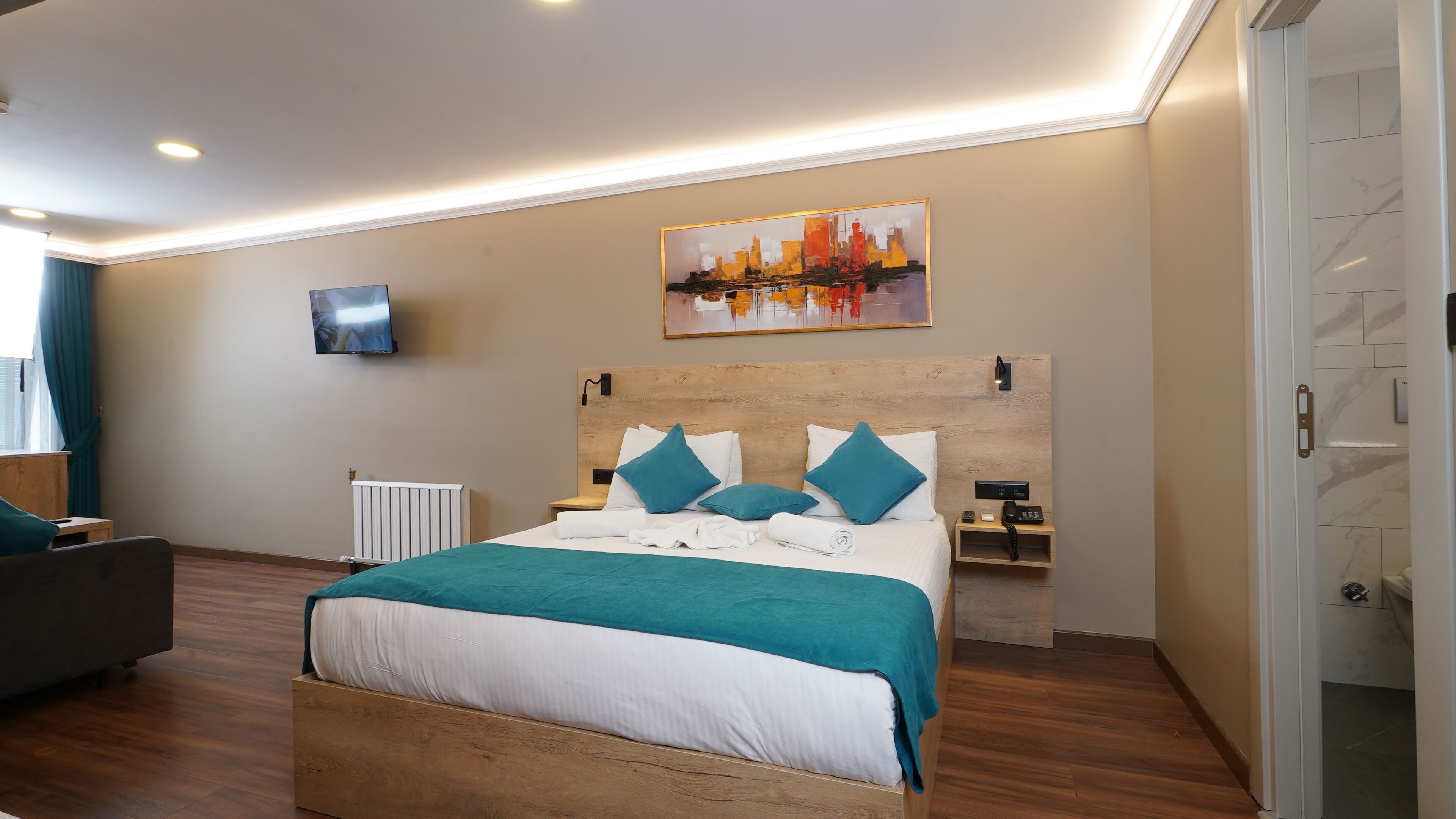 Una habitación de hotel moderna con una cama de madera, ropa de cama color turquesa, un televisor de pantalla plana, un cuadro y una mesita de noche con un teléfono.