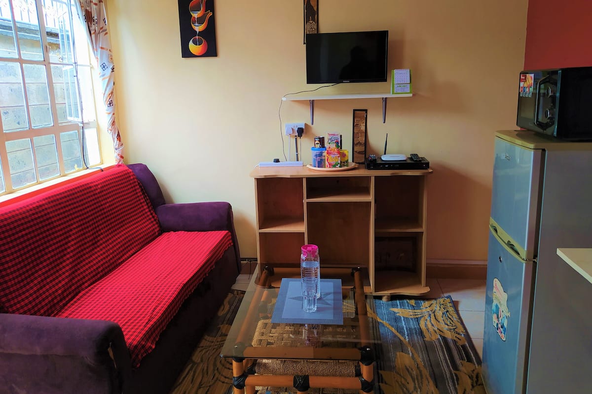 Ein gemütlicher Wohnraum mit Sofa, kleinem Tisch mit einem Glas, Fernseher, Kühlschrank und einem Fenster mit Vorhängen.