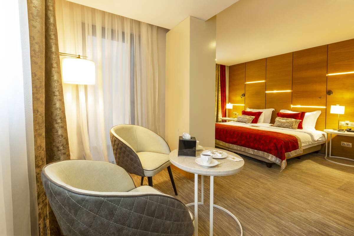 Een hotelkamer met een bed, twee stoelen, een klein tafeltje met een kopje en een pak tissues, en een lamp.